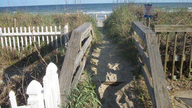 A sandy beach path leading to the ocean, flanked by a white picket fence and a wooden boardwalk. - Beautiful Efficiency Condo Across The Street from Beach. Great Pool & Deck Area. (North Myrtle Beach)