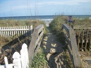 A sandy beach path leading to the ocean, flanked by a white picket fence and a wooden boardwalk.