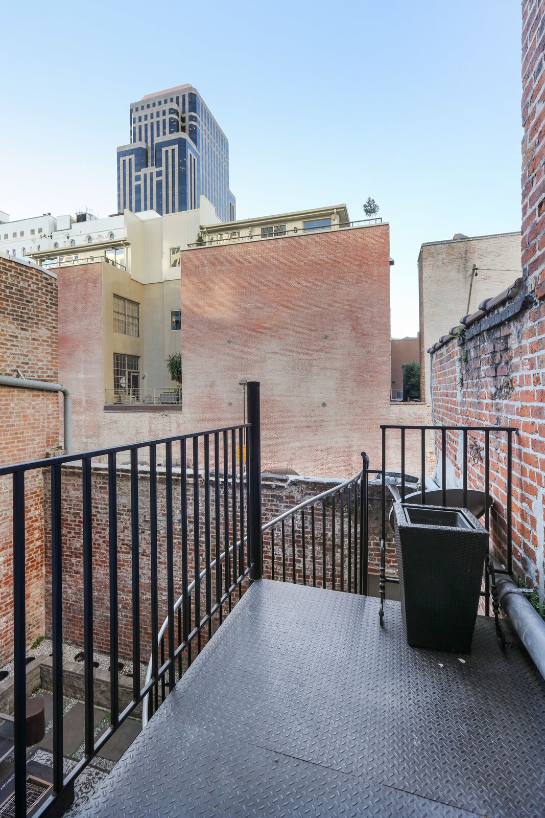Una terraza en la azotea con barandilla metálica, un basurero negro y vistas de edificios circundantes.