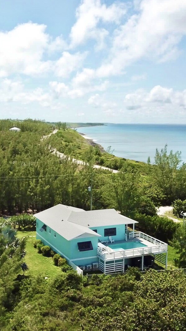 A two-story house with a pool on the second floor, surrounded by lush greenery and overlooking a beach.