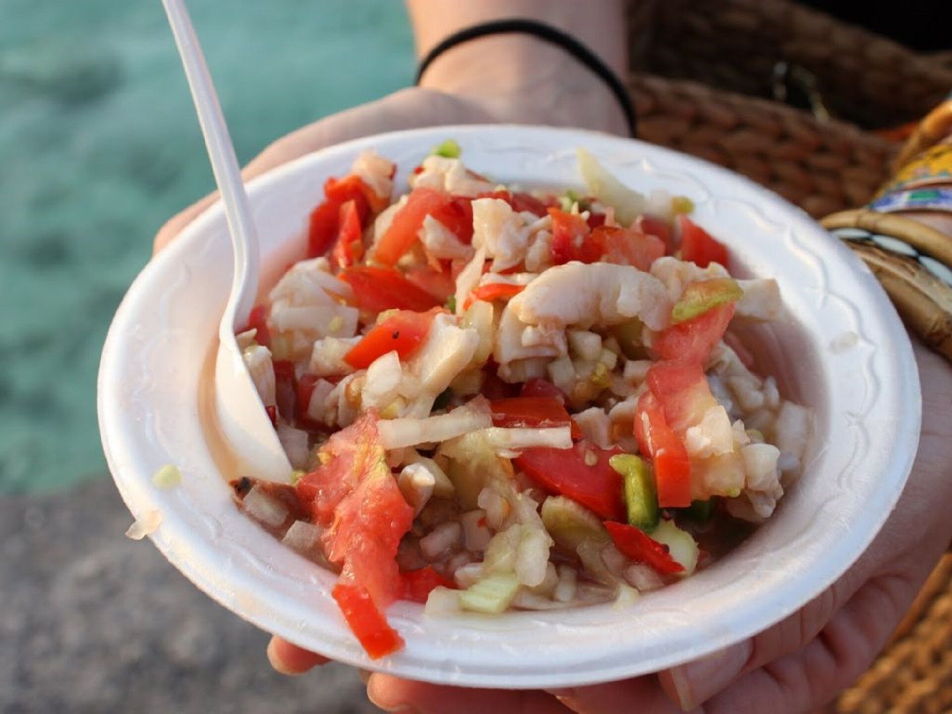 A person holding a white disposable bowl containing a seafood salad with visible ingredients like shrimp, tomatoes, and onions.
