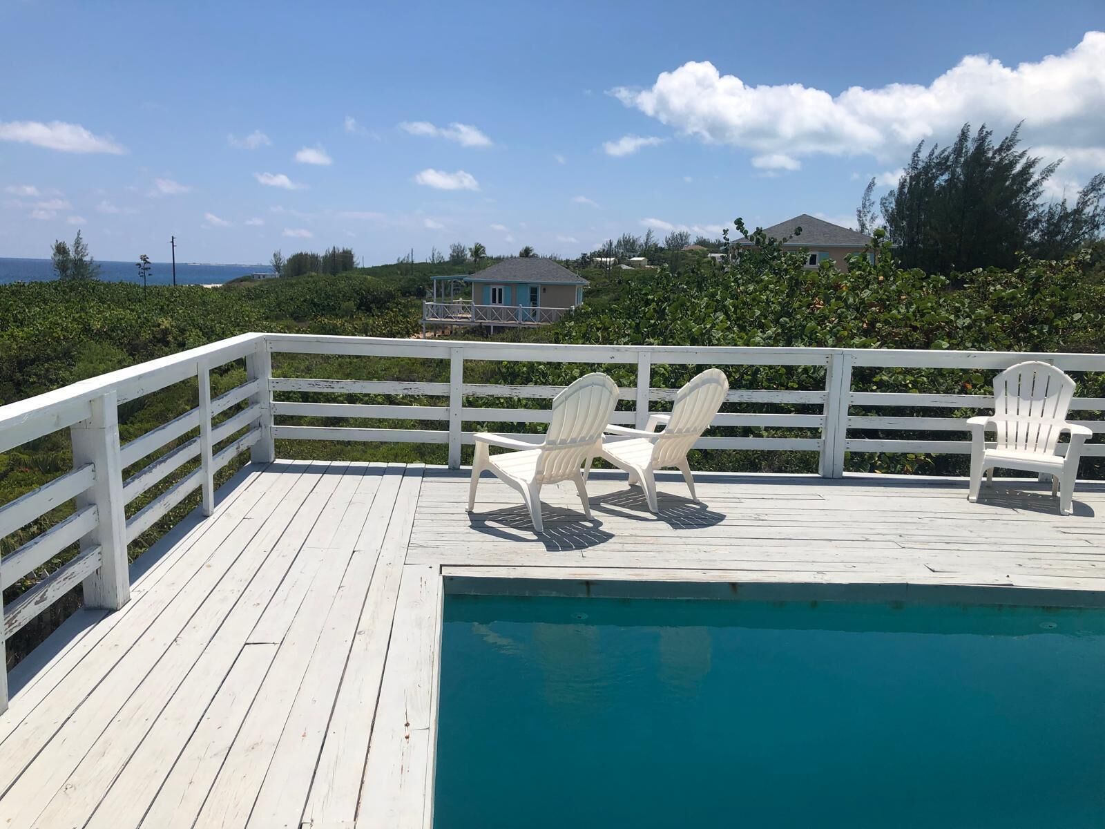 A wooden deck with a pool, two Adirondack chairs, and a view of a house and greenery.
