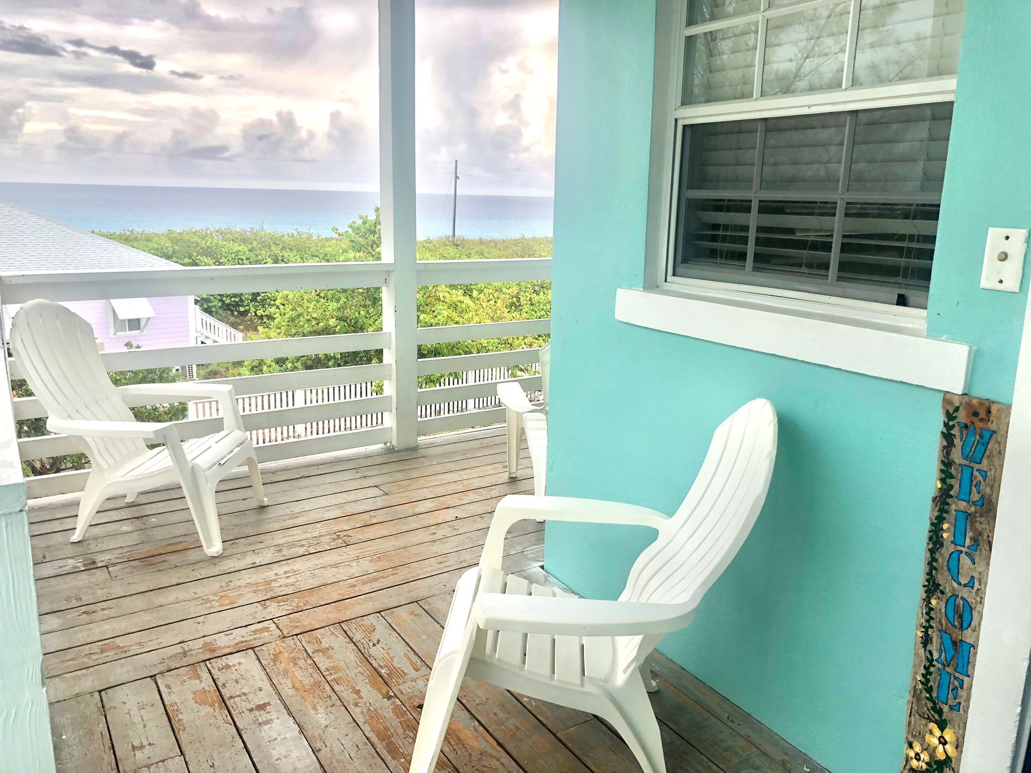 A wooden deck with two white plastic chairs, a turquoise wall, and a window with blinds.
