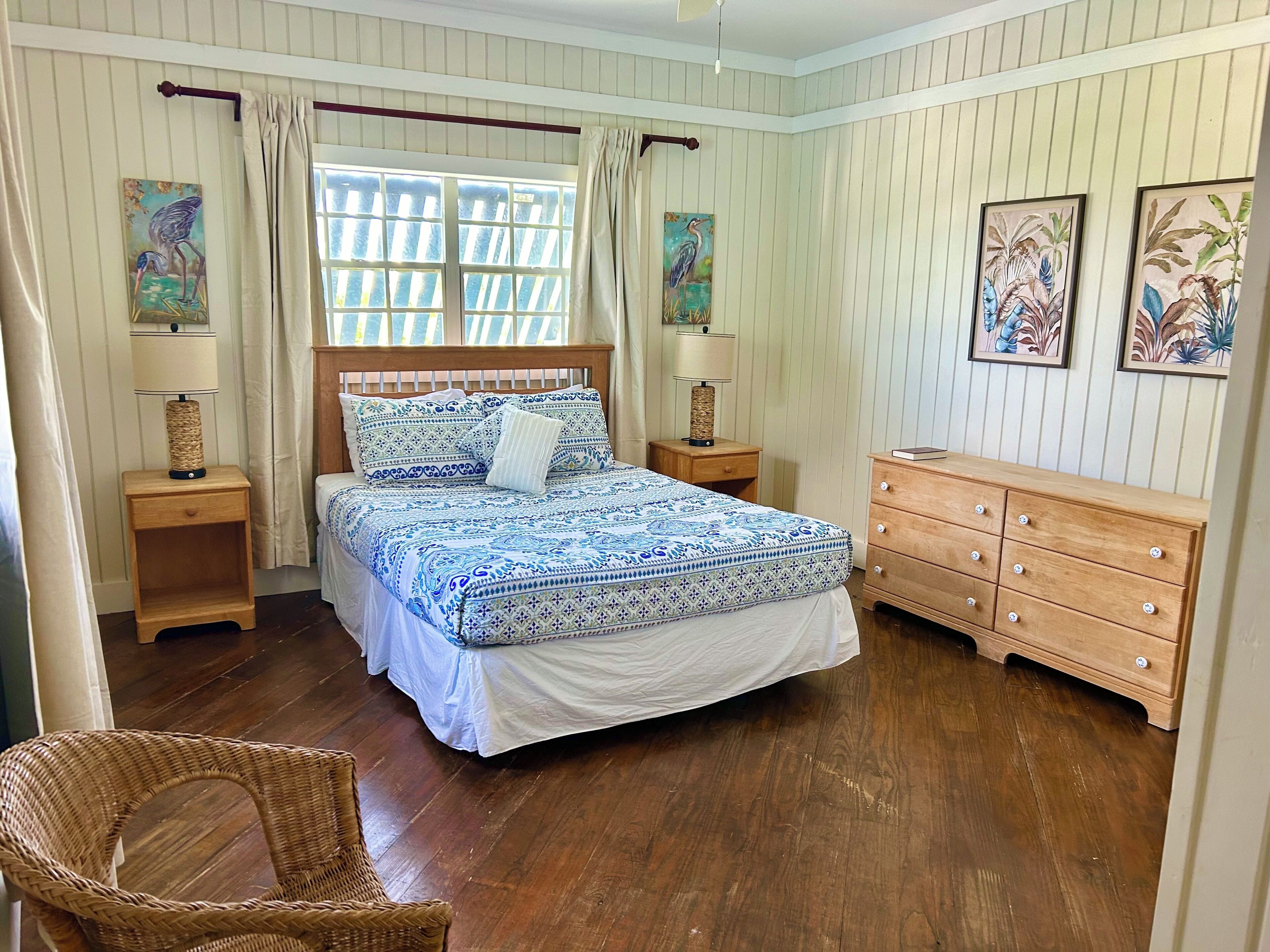 Un dormitorio con cama de madera, dos mesitas de noche, un aparador y una silla de mimbre.