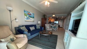 A living room with a blue sofa, beige armchair, and a coffee table. There is a painting on the wall, a ceiling fan, and a kitchen in the background. - Crystal Shores, Gulf Shores. Family Friendly Beachfront Condo w/ Pool,GYM & more (Gulf Shores)