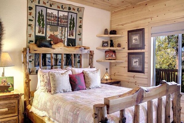 A cozy bedroom with a wooden bed frame, a quilted bedspread, and a wall decorated with animal prints. There are shelves with framed pictures and a window with a view of greenery.