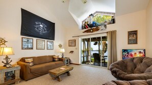 A living room with a brown sofa, a wooden coffee table, and a large black flag with a skull and crossbones design. There is a window with curtains, a framed picture on the wall, and a small table with a lamp. - Serenity by the Sea at Whitman Point. Gulf front and Screened Porches (Cedar Key)