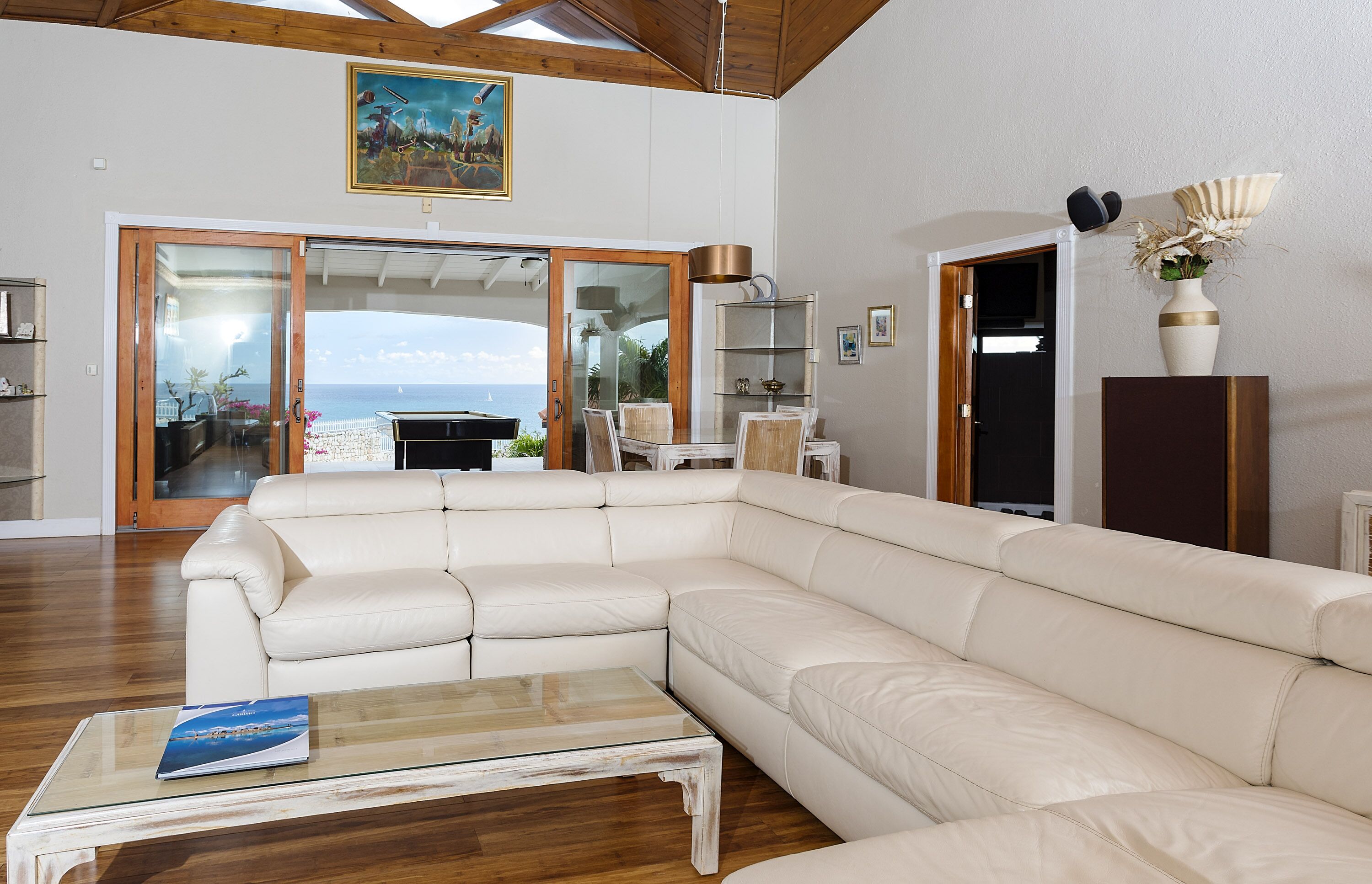 Een ruime woonkamer met een grote witte leren bank, een glazen salontafel en uitzicht op de oceaan door een schuifdeur.