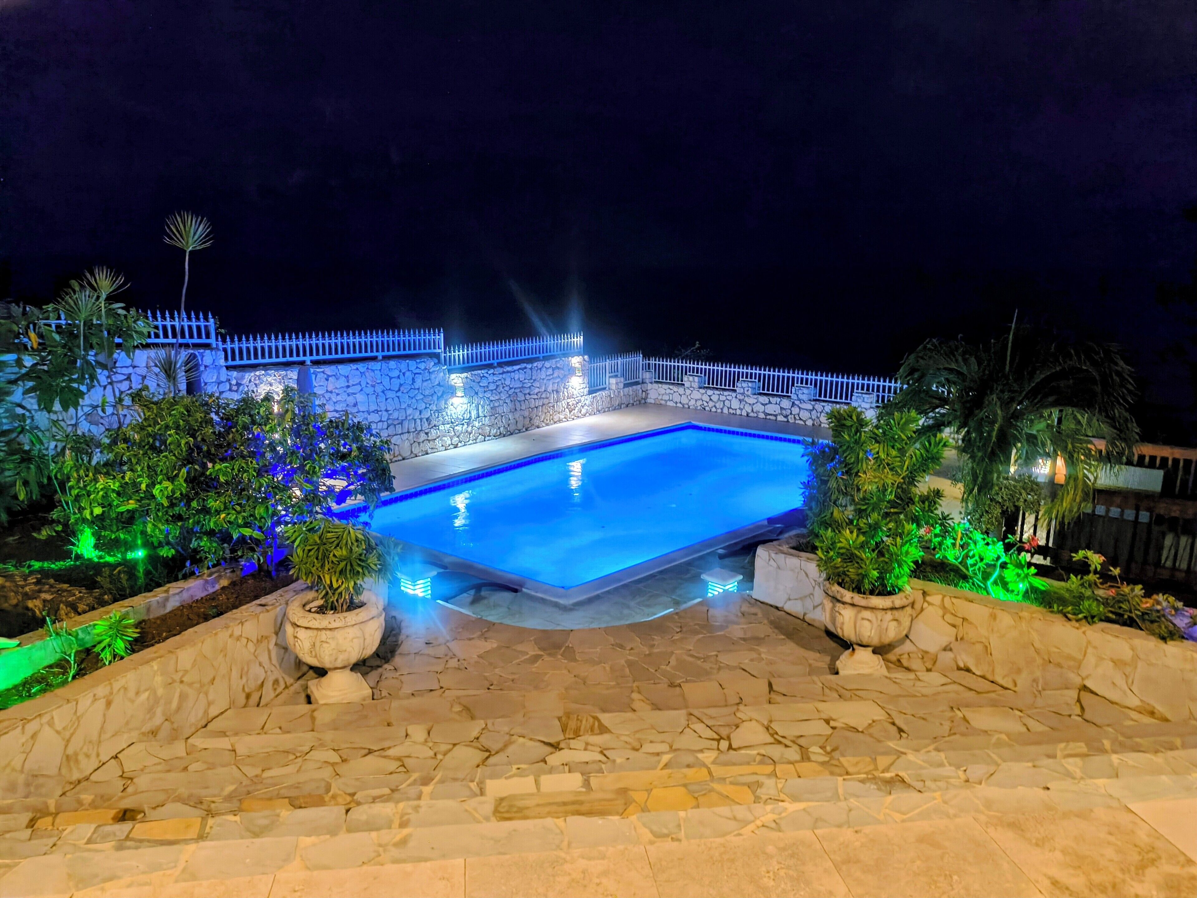 Une piscine aux murs en pierre et à la clôture blanche, entourée de plantes en pot et illuminée par des lumières bleues et vertes.