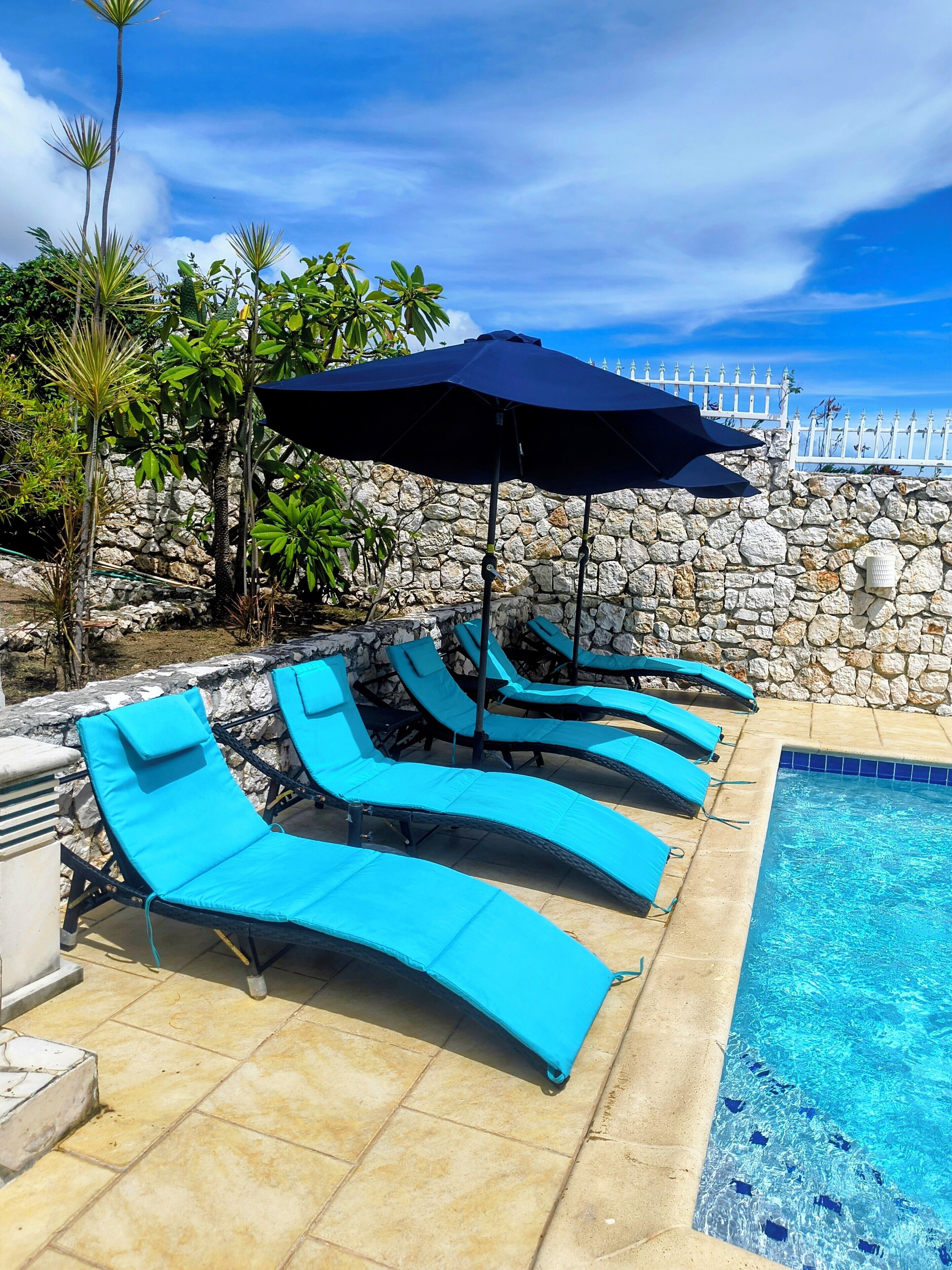 A poolside area with lounge chairs and an umbrella, surrounded by a stone wall and tropical plants.