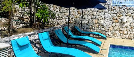 A poolside area with lounge chairs and an umbrella, surrounded by a stone wall and tropical plants.