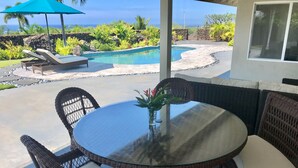 A glass-top table with wicker chairs, a small vase with flowers, and a view of a pool and ocean.