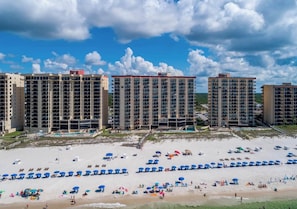 Un hotel frente a la playa con varios edificios de gran altura, una playa arenosa con sombrillas y un cielo despejado.