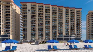 Ett högt hotell med flera balkonger, ett strandområde med solstolar och parasoller samt en klarblå himmel.