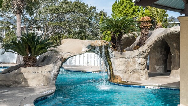 A swimming pool with a waterfall feature, surrounded by palm trees and rocks. - The Misty Breeze! Cabana Club, Ocean View, Golf Cart  (Fripp Island)