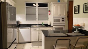 Una cocina con armarios blancos, encimera negra, microondas, lavavajillas y una ventana con persianas.