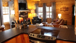 Un interior acogedor estilo cabaña con zona de cocina que incluye una estufa, mesa de comedor y rincón de estar con sofá y sillones.