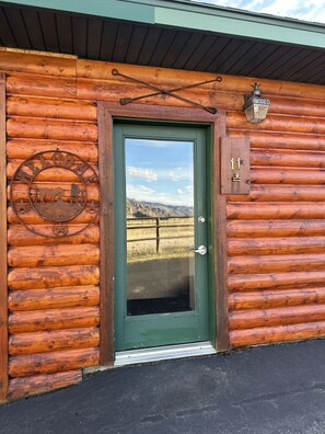 Une cabane en rondins avec une porte verte, une pancarte de bienvenue et une plaque numérotée.