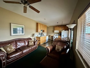 A living room with a brown leather sofa, a dining table, and a kitchen area with wooden cabinets and appliances.