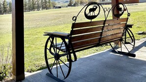 A bench with a metal frame and wooden slats, positioned under a roofed area with a view of a grassy field and mountains in the background. - Looking for an Alternative to a Big Box Motel?  We're it! (Gardiner)