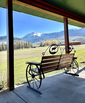 A bench with a metal frame and wooden slats, positioned under a roofed area with a view of a grassy field and mountains in the background.