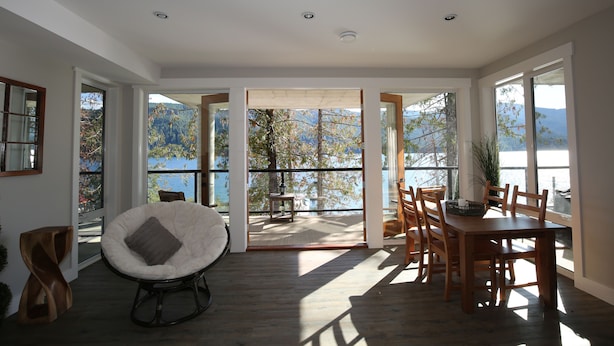 A spacious room with a wooden dining table and chairs, a white cushioned chair, and large windows offering a view of a lake and mountains.