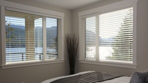 A bedroom with a bed, two windows with blinds, a vase with branches, and a view of mountains and buildings.