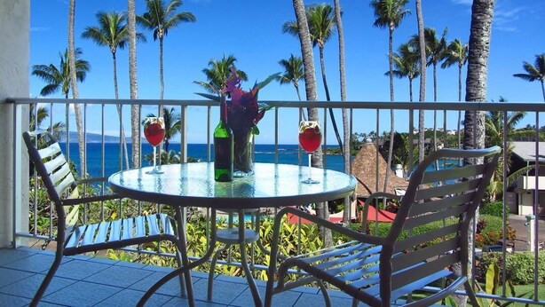 Ein Balkon mit einem runden Tisch und zwei Stühlen, der auf eine tropische Landschaft mit Palmen und blauem Himmel blickt.
