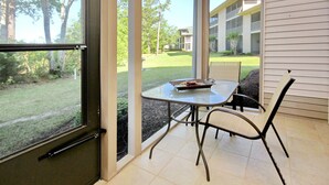A patio with a glass table and chairs, a small table with a bowl of fruit, and a view of a building and trees through a glass door.