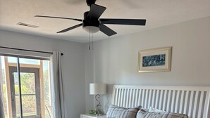 A bedroom with a bed, a ceiling fan, a nightstand with a lamp, a framed picture on the wall, and a window with curtains.