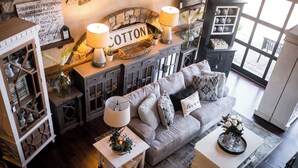 En hyggelig stue med en grå sofa, to hvide sengeborde og et sofabord. Der er dekorative lamper, et skilt med teksten "COTTON" og en pejs af sten.