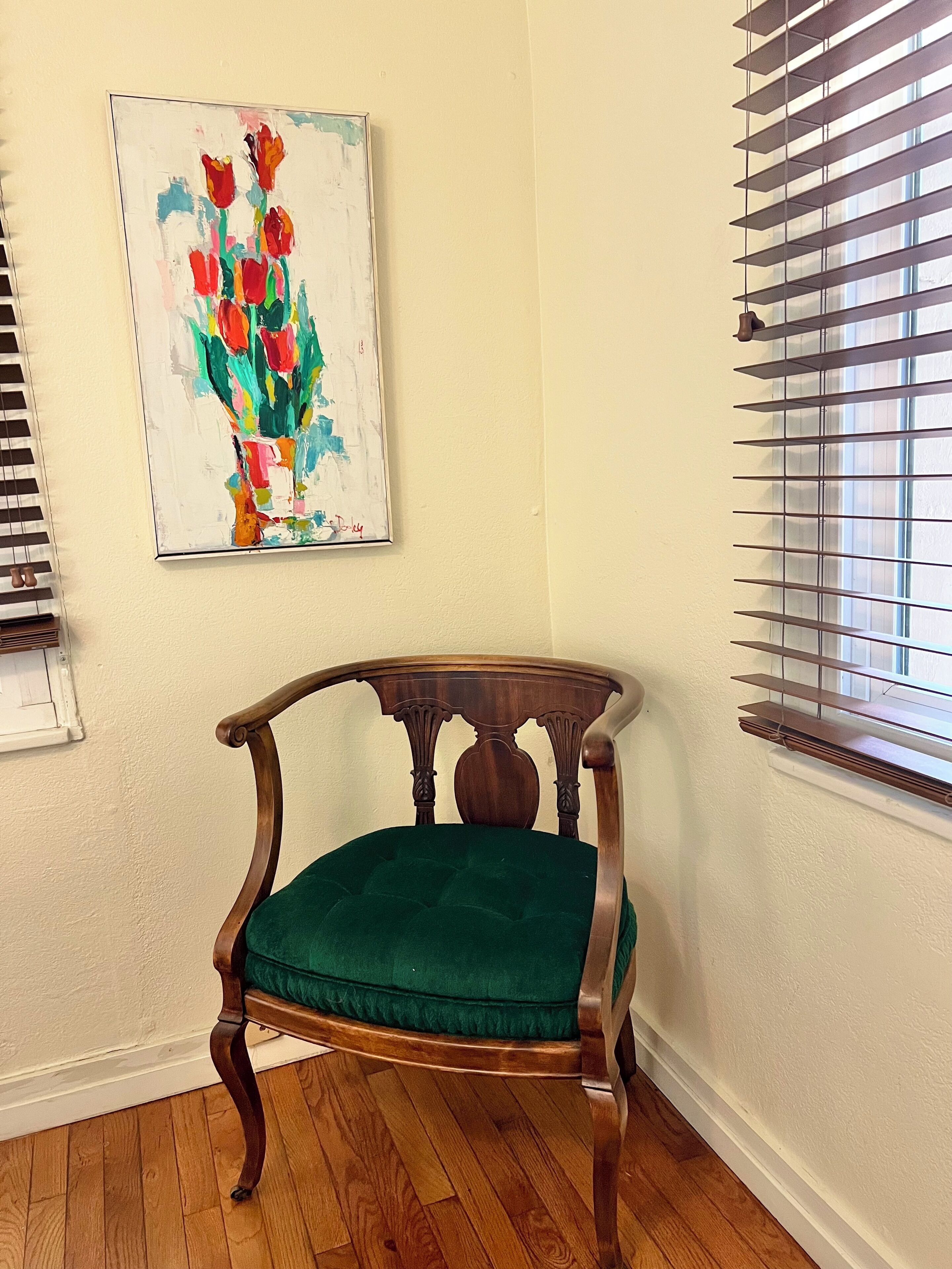 A corner room with a wooden chair, a painting of flowers, and a window with blinds.
