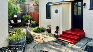 Ein kleines Haus mit roter Tür, Holzlattenzaun und einem Garten mit Blumentöpfen und einer Bank.