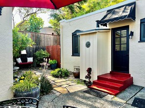 A small house with a red door, a wooden fence, and a garden with potted plants and a bench.