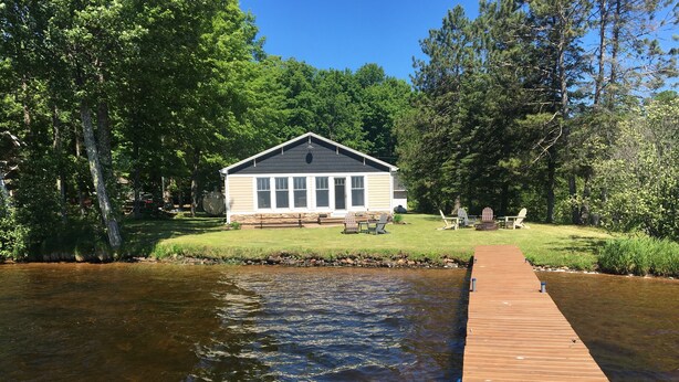 Eine Blockhütte am Seeufer mit einer anlegenden Pier, umgeben von Bäumen und einer Wiese mit Außenmöbeln.