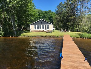 Una cabaña junto al lago con un muelle que se adentra en el agua, rodeada de árboles y un  área verde con mobiliario de exterior.