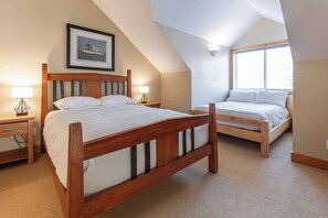 Une chambre avec un grand lit, un plus petit lit, une table de chevet avec une lampe, un tableau encadré au mur et une fenêtre.