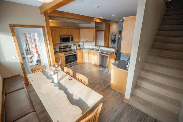 Una cocina moderna con armarios de madera, un comedor con mesa y sillas, y una escalera que sube a otro nivel.