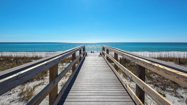 Een houten looppad leidt naar een strand met helderblauw water en een stralende zon.