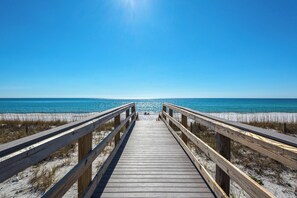 A wooden boardwalk leading to a beach with clear blue water and a bright sun in the sky.