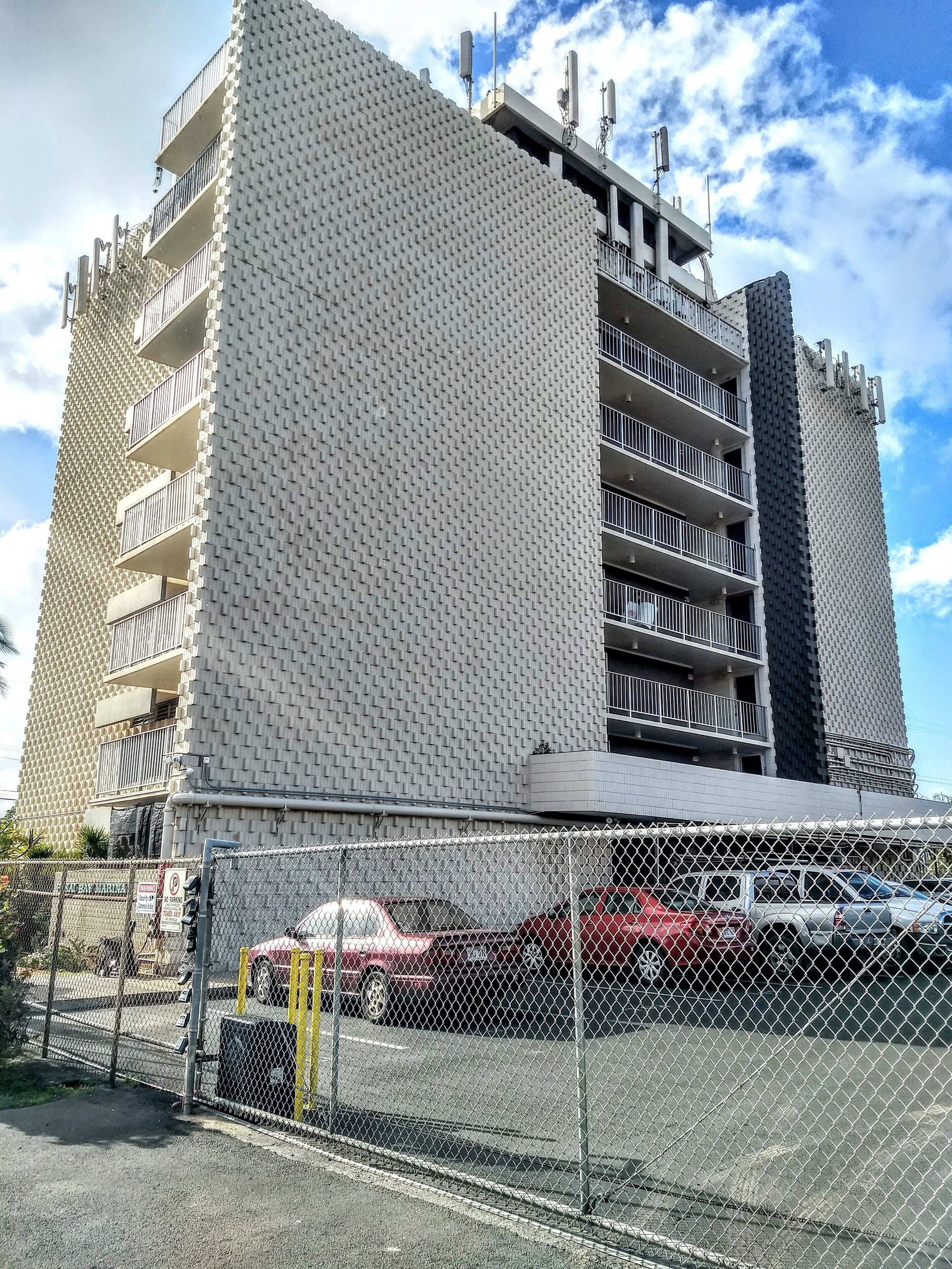 Un edificio de varios pisos con una fachada texturizada distintiva, balcones y un estacionamiento con cerca de alambre.