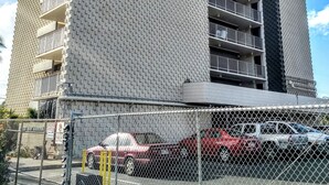 Un edificio a più piani con una facciata dalla texture particolare, balconi e un’area di parcheggio delimitata da una recinzione in rete metallica.