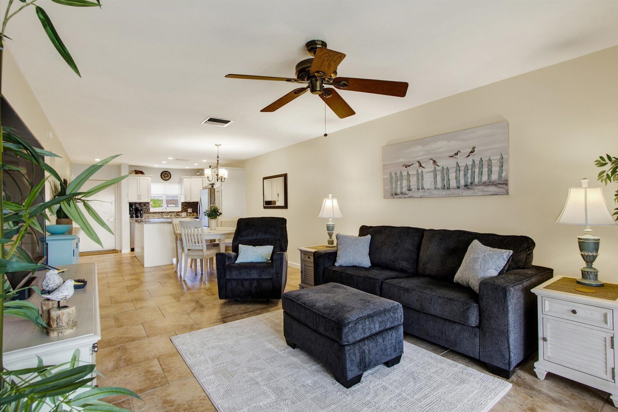A spacious living room with a dark grey sofa, matching ottoman, and a ceiling fan. A dining area with a table and chairs is visible in the background.