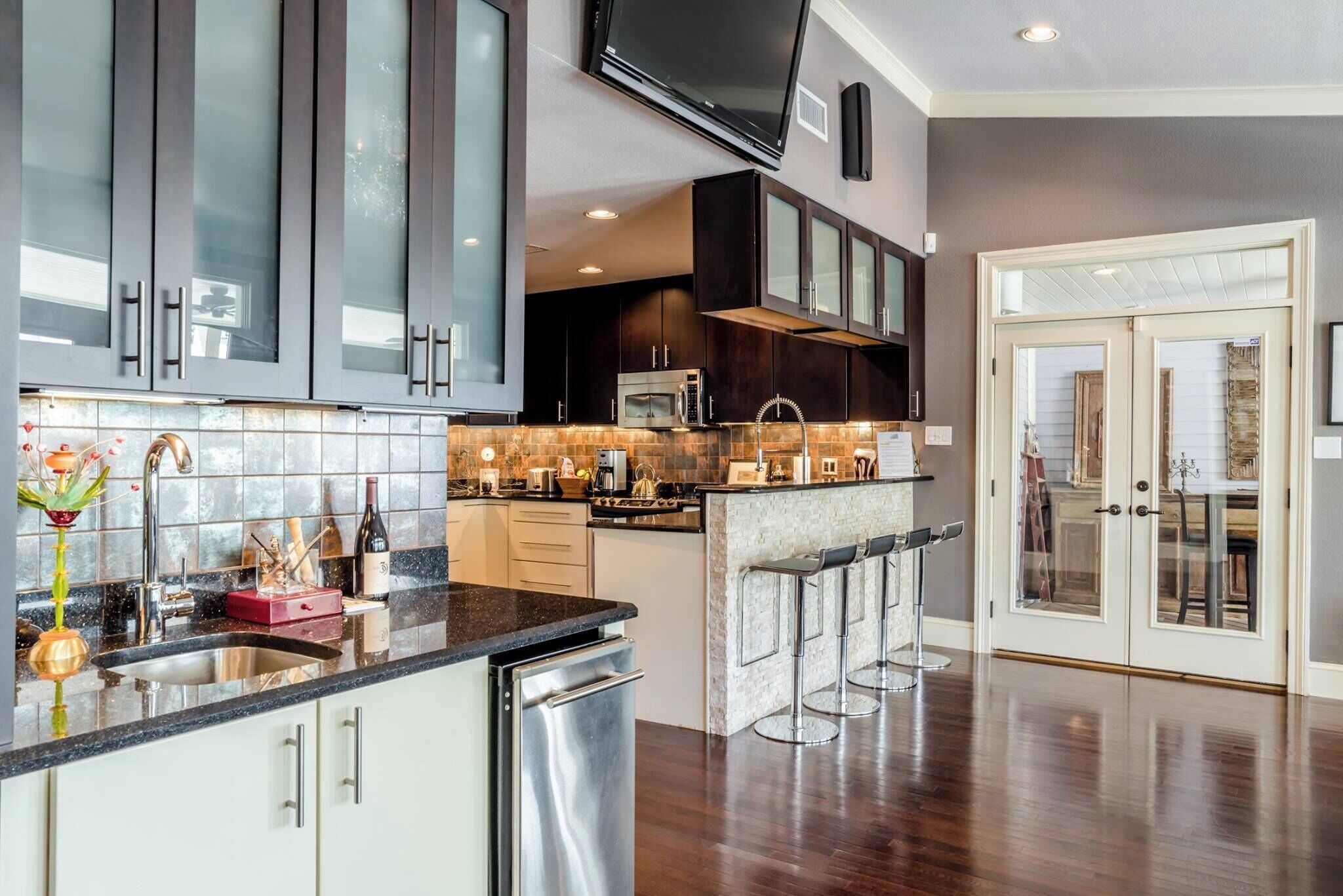 A modern kitchen with dark cabinets, a stainless steel dishwasher, a microwave, and a large TV mounted above the counter.