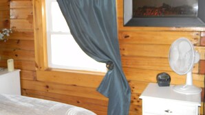 A wooden cabin room with a bed, a window with curtains, a wall-mounted heater, and a small white cabinet with a fan.
