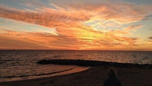 A beach with a sunset, a person sitting on the sand, and a rocky shoreline.