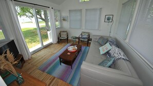 A spacious living room with a large sofa, a coffee table, and two chairs. There are large windows with blinds, a fireplace, and a view of the outdoors.