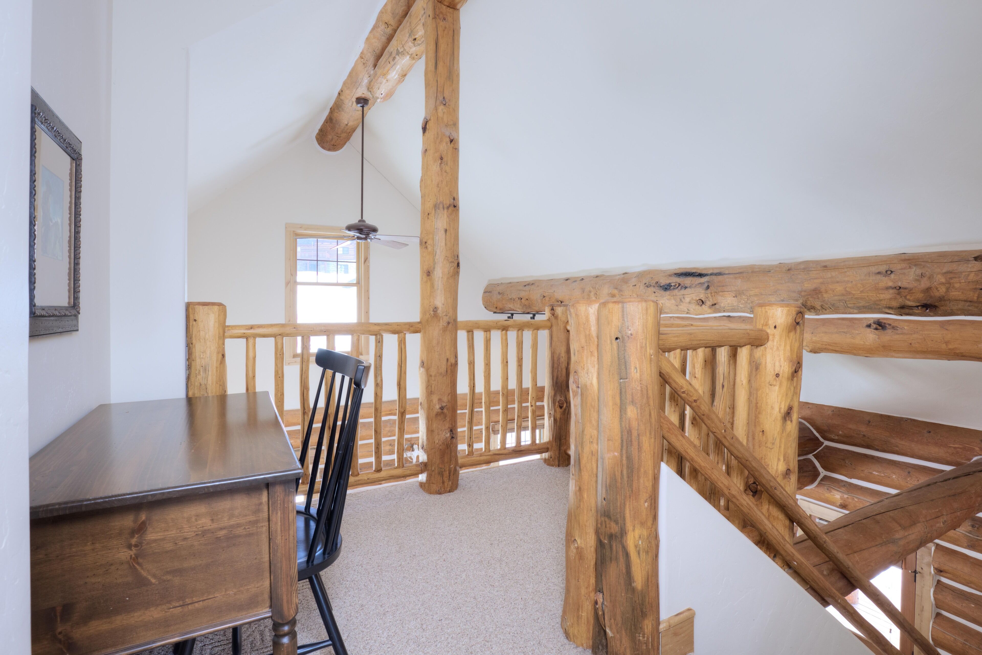 A wooden staircase with a railing, a wooden desk with a chair, and a ceiling fan in a room with white walls and a window.