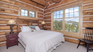 A log cabin bedroom with a large window, wooden walls, a bed with white bedding, two bedside tables with lamps, and a chair. - Ski-In/Ski-Out Powder Ridge Cabin In Beautiful Big Sky (Big Sky)