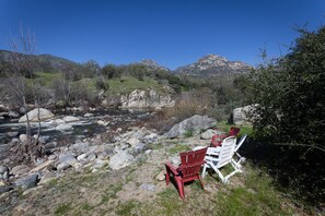 Un entorno junto al río con una mesa y dos sillas, rodeado de rocas y vegetación, y montañas al fondo.